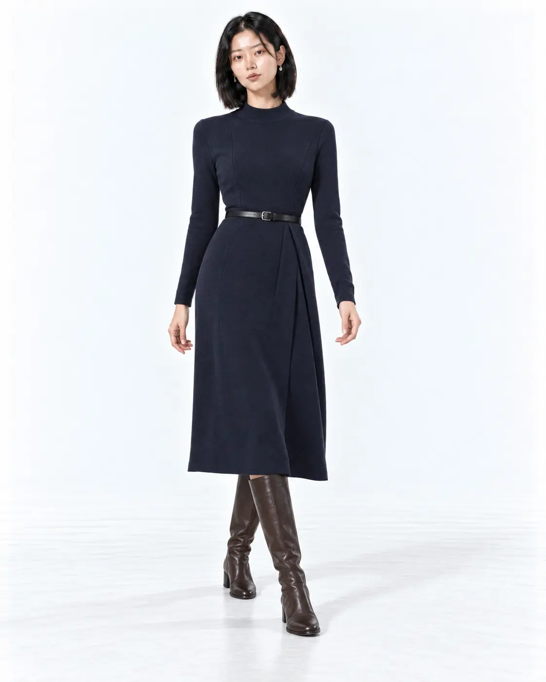 Navy Merino Knit Office Dress Woman wearing an ink navy merino midi dress with a slim belt and brown knee-high boots in a white studio.
