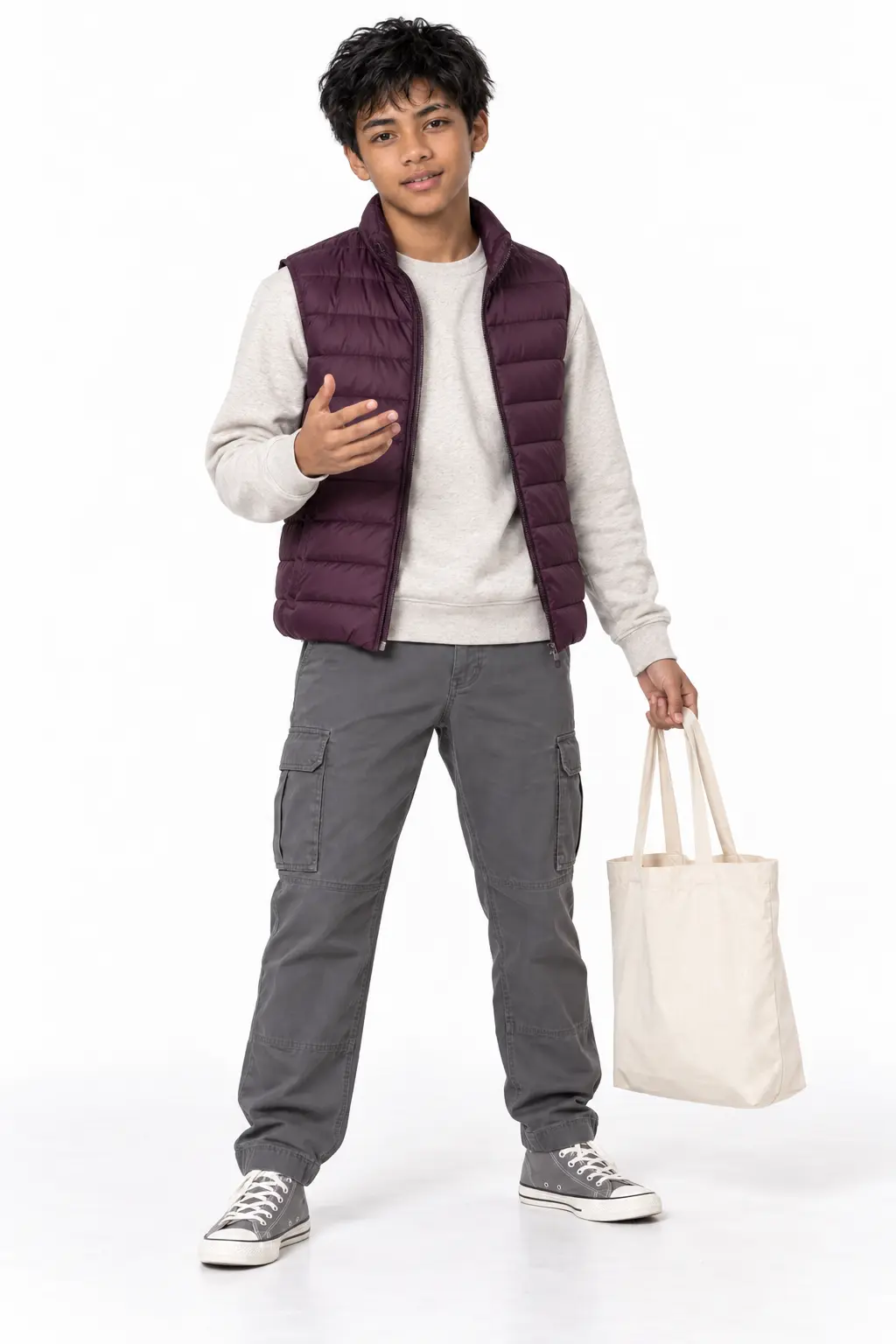 Teen Boy in Plum Puffer Vest and Grey Cargo Pants Holding Canvas Tote Bag Full-length studio portrait of a teenage boy wearing a sleeveless plum purple puffer vest over a light grey crewneck sweatshirt, charcoal cargo pants, and grey high-top sneakers while holding a cream canvas tote bag.