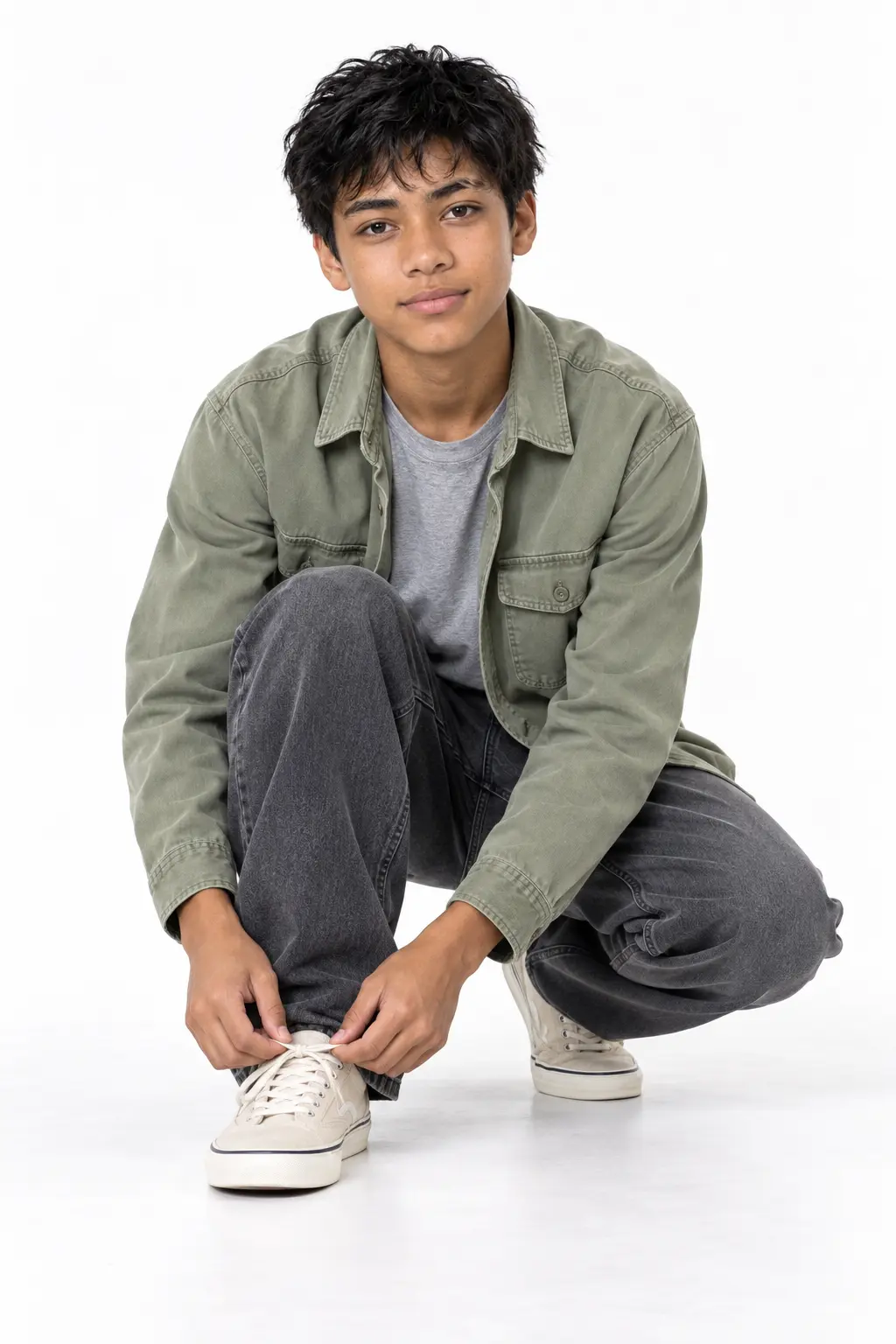 Teen Boy Crouching in Sage Green Utility Overshirt and Charcoal Denim Jeans A teenage boy crouching down to tie his cream sneakers, wearing a sage green button-up overshirt layered over a grey t-shirt and baggy charcoal grey jeans on a white studio background.